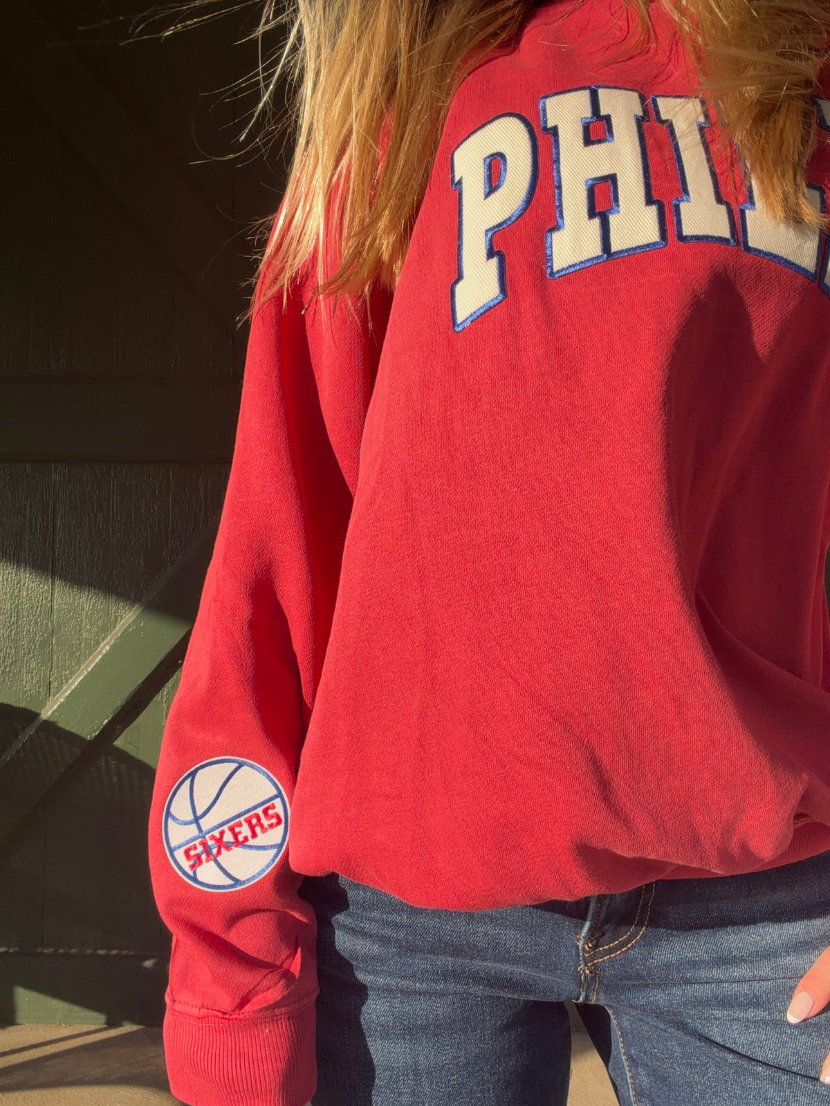 Red sweatshirt detail showing PHILLY lettering and a circular 76ers patch on the sleeve